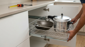 A close-up photograph of a stainless steel pull-out basket organizer holding pots and pans in a modern kitchen cabinet, illustrating efficient under-sink storage.
