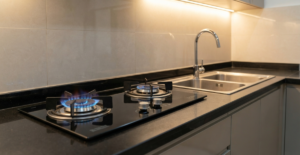 A premium kitchen setup in Bangladesh showcasing a built-in 2-burner tempered glass gas stove and a large stainless steel double-bowl sink set into a dark quartz countertop.