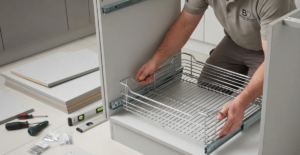 A close-up view showing a technician installing a stainless steel kitchen drawer basket system into a waterproof modular cabinet carcass during the installation process.