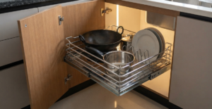 An open modern base cabinet in a Bangladeshi kitchen, revealing a heavy-duty stainless steel pull-out drawer basket organizing pots and pans, demonstrating modular storage solutions.